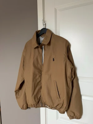 Brun bomberjacka med broderad logga - Snygg brun bomberjacka med klassisk krage och elastiska muddar. Jackan har två fickor framtill och en broderad logga på bröstet. Perfekt för dig som gillar en stilren och avslappnad look.