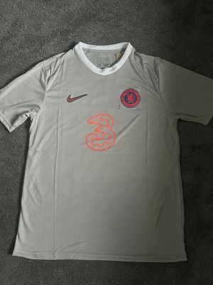 Chelsea grå matchtröja Nike - Chelsea FC matchtröja i ljusgrått med korta ärmar och vit krage. Tröjan har Nike-logga i vinrött, Chelsea-märke på bröstet och stor röd sponsorlogga i mitten. Tillverkad i lätt och ventilerande material, perfekt för fotbollsträning eller match.