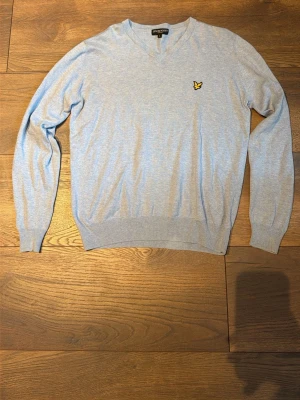Ljusblå stickad tröja Lyle & Scott - Ljusblå stickad tröja från Lyle & Scott med V-ringning och deras klassiska gula logga på bröstet. Tröjan har ribbade muddar vid ärmslut och nederkant. Perfekt för dig som gillar stilrena och bekväma plagg. STORLEKN ÄR S/M, skulle säga närmare M än S. 
