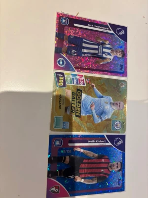 Premier League samlarkort 3-pack - Tre glittriga samlarkort från Premier League med spelarna Jack Hinshelwood, Haaland (Golden Baller) och Justin Kluivert. Kort i olika färger: rosa, guld och blå, med spelarnas namn, position och klubbmärke tydligt tryckta. Perfekt för dig som gillar fotboll och att samla kort. Samt att Justin kluivert och Jack Hinshelwood är numererade Justin är till 150 och Jack är till 399