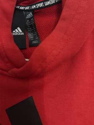Röd hoodie från Adidas med huva - Snygg röd hoodie från Adidas med klassisk huva och svart Adidas-logga på bröstet. Tillverkad i mjukt material som känns skönt mot huden. Perfekt för dig som gillar sportig och avslappnad stil.