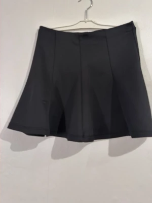 Svart utsvängd kortkjol - Snygg svart kortkjol med utsvängd passform och stilren design. Kjolen har en enkel look utan synliga detaljer och passar perfekt till många olika outfits. Tillverkad i ett mjukt och följsamt material som ger en bekväm känsla.