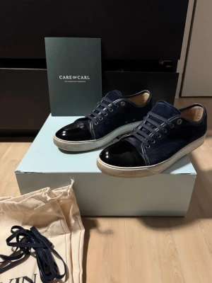 Lanvin skor - Snygga marinblå sneakers från Lanvin med glansiga lackdetaljer på tån och klassisk vit sula. Skorna har marinblå skosnören och metallöljetter, samt en ovandel i mocka och läder. Perfekta för dig som vill ha en stilren och exklusiv look. Köpta från CareofCarl, alla tillbehör ingår såklart. Liten skada på ena skon, se sista bild. Prisförlsag och frågor är varmt välkomna! 