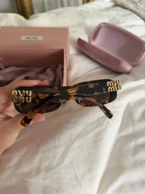 Miu Miu solglasögon - Superfina Miu Miu-solglasögon i Honey Havana/tortoise med guldfärgade Miu Miu-detaljer. Unisexmodell som inte säljs i ordinarie Miu Miu-sortiment längre.  Modell: SMU 08Z Färgkod: VAU-06B Storlek: 53-20-140 Nypris ca 5 000 kr.  Fint begagnat skick. Ena skalmen är något lös och skulle behöva dras åt/justeras hos optiker, därav priset. Det finns även några mycket små ytliga repor på glasen som endast syns om man granskar dem i starkt ljus/solljus – ingenting som påverkar sikten när de används 🩷