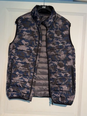 Camouflagefärgad dunväst med dragkedja - Snygg dunväst i blått, grått och svart camouflage-mönster. Västen har hög krage, två fickor med dragkedja och quiltad design. Knappt använd, inga tecken på defekter. Hör av er vid funderingar.