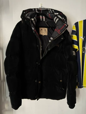 Burberry pufferjacka - Säljer en svart pufferjacka från Burberry i storlek M. Jackan har en klassisk Burberry-rutig huva i grått, rött och vitt, dragkedja och knappar framtill samt två fickor med lock. Perfekt för kalla vinterdagar och riktigt snygg streetstil.