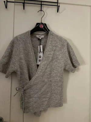 Grå omlottstickad topp kort ärm - Säljer en ljusgrå stickad omlottopp med korta ärmar och knytband i midjan. Toppen har en djup V-ringning och är gjord i mjukt, tunt stickat material. Perfekt för lager på lager och enkel att styla till olika outfits.