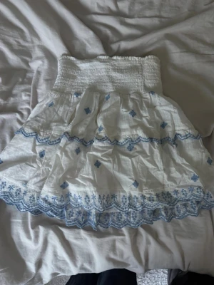 Vit kortkjol med blå broderier - Söt vit kortkjol med blå broderade detaljer och volangkant. Kjolen har en bred, smockad midja som ger en bekväm passform och ett luftigt, somrigt material. Perfekt för varma dagar och enkel att matcha med olika toppar.