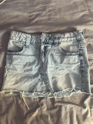 Ljusblå jeanskjol med rå kant - Trendig ljusblå jeanskjol i kort modell med slitna detaljer och rå, fransig kant nedtill. Kjolen har klassiska fickor fram och hällor för bälte. Perfekt för en avslappnad och cool stil.
