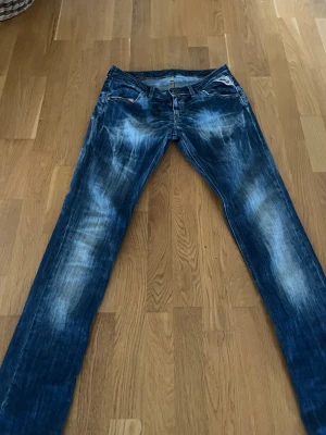 Replay jeans - Säljer ett par blå jeans med slitningar och tvättade detaljer. Jeansen har raka ben, klassisk femficksmodell och låg midja. Materialet är denim och de har en cool, avslappnad look med ljusa partier framtill och bakfickor med broderad text. Storlek 29