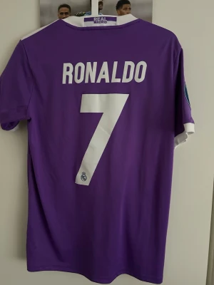 Lila Real Madrid Ronaldo matchtröja - Säljer en lila Real Madrid matchtröja med Ronaldo och nummer 7 på ryggen. Tröjan har vita detaljer, klubbmärke, Adidas-logga och Fly Emirates-tryck på bröstet samt Champions League-patchar på ärmarna. Perfekt för dig som älskar fotboll och vill sticka ut på planen.