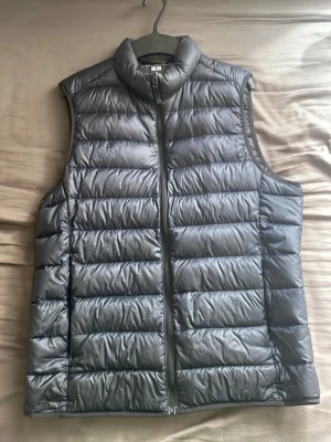 Svart dunväst med dragkedja från Uniqlo - Snygg svart dunväst från Uniqlo med hög krage och dragkedja framtill. Västen har en quiltad design med horisontella sömmar och är tillverkad i ett glansigt material som ger en modern look. Perfekt att slänga över en hoodie eller tröja för extra värme.