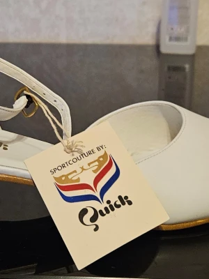 Vita pumps från Quick med låg klack - Snygga vita pumps från Quick med låg klack och öppen häl. Skorna har ett elegant band runt hälen med svart och vitt mönster samt gulddetalj. Perfekta för dig som gillar retrostil och sportig elegans.