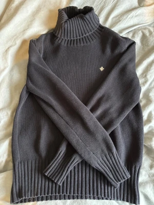 Morris Polo tröja - Mysig mörkgrå stickad polotröja med ribbad krage, ärmslut och nederkant. Tröjan har en liten broderad detalj på bröstet och är perfekt för lager på lager. Klassisk och enkel design som passar till det mesta.