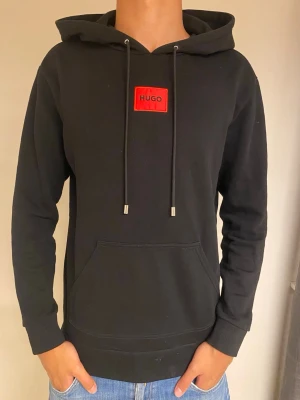 Svart hoodie från Hugo med röd logga - Svart hoodie från Hugo med röd logga på bröstet. Klassisk huva med dragsko och stor magficka framtill. Tillverkad i mjuk bomull och har långa ärmar med ribbade muddar. Perfekt för en avslappnad och stilren look.