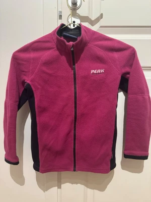 Rosa fleecejacka Peak Performance - Mysig rosa fleecejacka från Peak Performance med svarta detaljer längs sidorna och ärmsluten. Jackan har hel dragkedja framtill och hög krage. Perfekt för kyliga dagar och enkel att matcha med andra plagg.