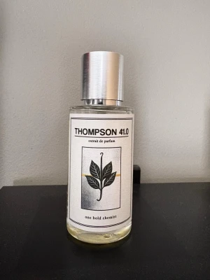 Thompson 41.0 parfym - En stilren parfymflaska med silverfärgat lock och transparent glas. Etiketten har en minimalistisk design med ett bladmotiv och texten 'THOMPSON 41.0' samt 'one bold chemist'. Innehållet är ljusgult och flaskan har en modern, enkel form ungefär 90% kvar av flaskan.