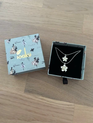 Halsband från Ioaku - Säljer ett super sött halsband från Ioaku med två blommiga hängen i silverfärg. Kommer i originalask. 