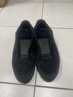 Svarta slip-on sneakers från Axel Arigato - Stilrena svarta slip-on sneakers från Axel Arigato i mocka med elastiska band över vristen. Skorna har en låg silhuett och diskret logga på sidan. Perfekta för dig som gillar minimalistisk och clean stil.