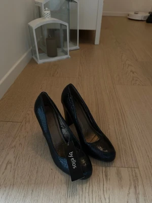 By Ellos klackskor – svarta – strl 40 🖤 - Snygga svarta pumps från by Ellos med trendigt ormmönstrad yta. Skorna har rundad tå och hög klack som ger en elegant siluett. Materialet ser ut som konstläder med glansig finish. Perfekta för att lyfta vilken outfit som helst.