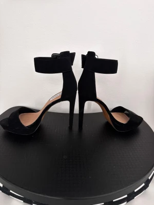 Svarta klackpumps från Steve Madden - Snygga svarta pumps med hög klack från Steve Madden. Skorna har öppen tå, bred rem över tårna och en bred ankelrem med spänne. Tillverkade i mockaliknande material och har en elegant siluett som ger en trendig look.