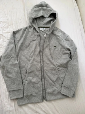 Grå zip hoodie från Lacoste - Snygg grå hoodie från Lacoste med dragkedja framtill, två fickor och klassisk huva med dragsko. Ikoniska gröna krokodilloggan på bröstet. Tillverkad i mjuk bomullsblandning som känns skön mot huden. Perfekt för en chill och stilren look.