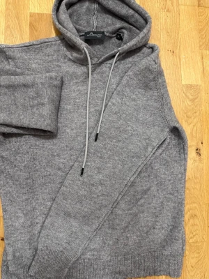 Grå hoodie från Montezemolo Trend  - Mysig grå hoodie från Montezemolo Trend med huva och dragsnören. Tillverkad i mjukt stickat material som känns skönt mot huden. Klassisk passform med ribbade muddar vid ärmslut och nederkant. Perfekt för en chill och avslappnad stil.