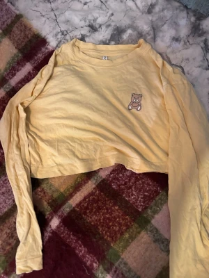 Gul cropped pyjamaströja med björn - Söt gul långärmad pyjamaströja i croppad modell med ett litet björnmärke på bröstet. Tillverkad i mjukt bomullstyg, perfekt för mysiga kvällar hemma. Enkel och avslappnad stil som passar perfekt till loungewear.