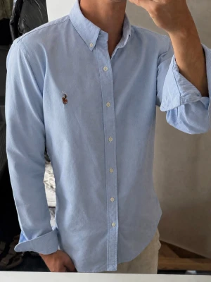 Ljusblå skjorta från Ralph Lauren - Klassisk ljusblå skjorta från Ralph Lauren med button-down krage och broderad logga på bröstet. Skjortan har långa ärmar, vita knappar och är tillverkad i ett mjukt bomullstyg. Perfekt för en clean och stilren look.