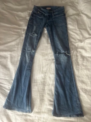 Blå utsvängda jeans med slitningar - Säljer ett par blå jeans med utsvängda ben och slitningar på båda knäna. Det finns en liten slitning längst ner på ena benet men annars är dom i nytt skick!