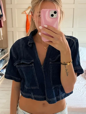 Mörkblå croppad jeansskjorta - Säljer en mörkblå croppad jeansskjorta med korta ärmar och två stora bröstfickor. Skjortan har en djup v-ringning och markerade sömmar som ger en cool look. Perfekt till jeans eller kjol för en avslappnad stil.