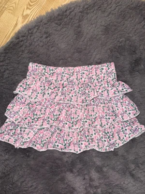 Rosa blommig volangkjol - Supersöt kort kjol med rosa blommigt mönster och flera volanger. Kjolen har elastisk midja och är perfekt för dig som gillar en romantisk och lekfull stil. Materialet känns lätt och luftigt, vilket gör den skön att bära under varma dagar. Barnstorlek 158-164.