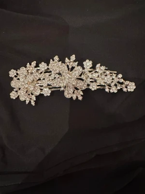 Glittrig hårklämma med blommor - Säljer en superfin hårklämma i silverfärg med gnistrande strass och detaljerade blommor. Klämman har en elegant och lyxig design som verkligen sticker ut. Perfekt för att ge frisyren det lilla extra och skapa en glamorös look. Klämman är 10,5 cm lång!