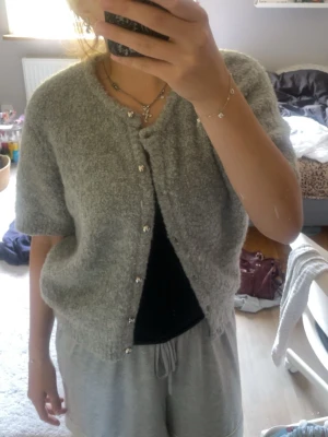Grå fluffig kofta med kort ärm - Mysig grå kofta med korta ärmar och fluffig textur. Koftan har fina blommiga knappar framtill och en avslappnad passform. Perfekt att slänga över en topp för en chill look.