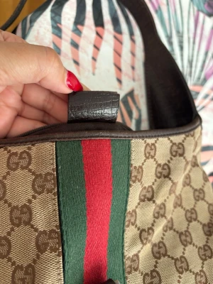 Gucci beige handväska med GG-mönster - Snygg beige handväska från Gucci med klassiskt GG-mönster och bred grön-röd rand framtill. Väskan har mörkbruna läderdetaljer och axelrem. Perfekt för dig som gillar tidlös design och vill ha en ikonisk accessoar.