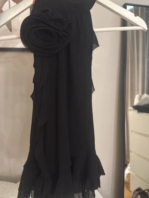 Svart halterneck topp - Snygg svart ärmlös topp med stor dekorativ blomdetalj på axeln och volangkant nedtill. Tillverkad i ett tunt, lätt och flygigt material som ger en elegant look. Perfekt för dig som vill sticka ut lite extra.