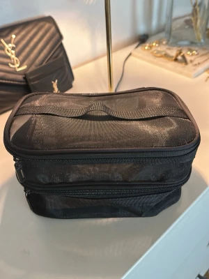 Svart mesh necessär med dragkedja - Praktisk svart necessär i meshmaterial med två separata fack och dragkedjor. Perfekt för att organisera smink, hudvård eller småprylar. Väskan har en rektangulär form och ett handtag på toppen för enkel transport.