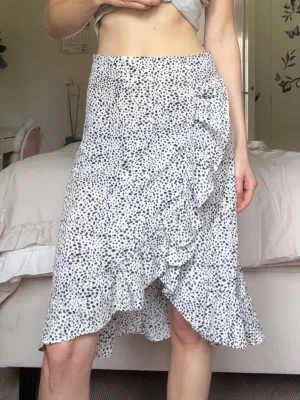 Vacker Vit prickig omlottkjol med volanger - Supersöt vit omlottkjol med svarta prickar (polkadot) och volangdetaljer längs kanten! Kjolen har en asymmetrisk nederkant och är tillverkad i ett lätt, flowy material som ger en luftig känsla🩷Den är längre bak och kortare framtill. Har även en dragkedja på sidan för lätt av&på tagning❤️Perfekt för dig som gillar en feminin och trendig stil, så bra plagg som passar till allt! Mått: 33cm midja, 66cm total bakre längd. Passar XS, inga defekter✨Hör av vid frågor eller bud🤍