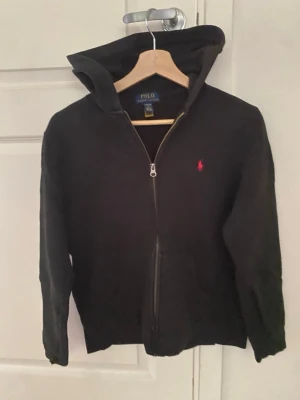 Svart hoodie från Polo Ralph Lauren - Svart hoodie med dragkedja från Polo Ralph Lauren. Klassisk design med huva, storlek (L14-16)