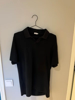 Svart pikétröja från Filippa K - Klassisk svart pikétröja från Filippa K i storlek L. Tröjan har krage och korta ärmar, perfekt för en clean och stilren look. Tillverkad i mjukt material som känns skönt mot huden.