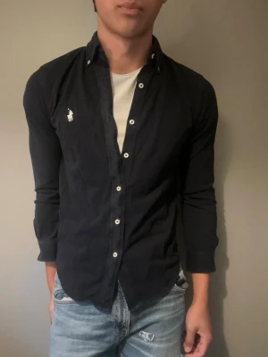 Ralph lauren skjorta - Snygg svart skjorta med vita knappar och klassisk krage. Skjortan har lång ärm och en vit broderad logga på bröstet. Perfekt till jeans eller chinos för en clean look.