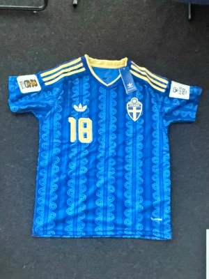 Sveriges Bortaställ 2026 (Ayari) - Säljer en blå Sverige fotbollströja från Adidas med gula detaljer och nummer 18. Tröjan har korta ärmar, gul krage och tre gula ränder på axlarna. Sveriges landslagsemblem på bröstet och namnet Ayari på ryggen. Perfekt för match eller träning.