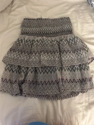 Missoni mönstrad kortkjol från KappAhl - Säljer en kortkjol från KappAhl med zigzag-mönster i svart, vitt, lila och grönt. Kjolen har en bred, smockad midja och dubbla volanger som ger en snygg och lekfull look. Perfekt för dig som gillar mönstrade plagg!