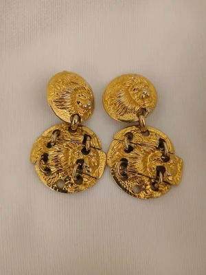 Guldpläterade clips - versace leo - Unika örhängen i guldpläterad metall med tvådelad design. Översta delen är rund med reliefmönster av ett lejonhuvud, och undre delen består av en större cirkel med samma motiv och länkade detaljer. Clips. En rolig anekdot/histoia kanske, dom har fått förgylla 3 talet tjejer på nobelfesten och premiären i cannes.