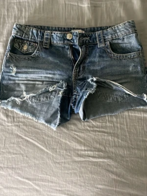 Blå jeansshorts med fransar - Säljer ett par blå jeansshorts med slitna detaljer och fransig kant. Shortsen har klassisk femficksdesign, bälteshällor och knappgylf. Perfekta för sommaren och ger en avslappnad vibe.