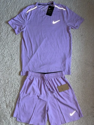 Lila träningsset från Nike - Snyggt lila träningsset från Nike med t-shirt och shorts. Båda plaggen har reflekterande Nike-logga och är gjorda i ett lätt och svalt material, perfekt för träning. T-shirten har korta ärmar och shortsen har resår i midjan.