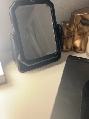 Svart sminkspegel med ställ - Snygg sminkspegel i svart plast med rektangulär form och lätt avfasade hörn. Spegeln står på ett stabilt ställ som gör det enkelt att vinkla den efter behov. Perfekt storlek för sminkbordet eller skrivbordet.