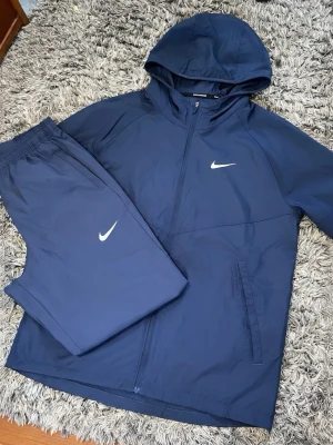 Marinblått Nike träningsset med huva - Snyggt marinblått träningsset från Nike med matchande jacka och byxor. Jackan har huva, dragkedja och två fickor, medan byxorna har resår i midjan. Båda plaggen har Nike-logga i vitt. Perfekt för löpning eller gymmet.