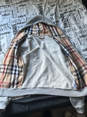 Grå hoodie med dragkedja och mönstrat foder från burbery - Snygg grå hoodie med dragkedja och fickor framtill. Insidan och huvan har det klassiska rutiga mönstret i beige, svart, vitt och rött. Hoodien har ribbade muddar och känns riktigt mjuk och skön. Perfekt för dig som gillar stilrena plagg med en twist.