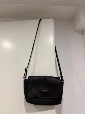 Svart axelväska från Björn Borg - Snygg svart axelväska från Björn Borg i konstläder med justerbar axelrem. Väskan har ett stilrent och enkelt utseende med metallogo framtill och lockstängning. Perfekt storlek för det viktigaste och passar till många olika outfits.